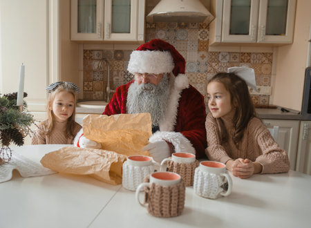 Santa Claus reads a Christmas letter with two adorable girls in a cozy kitchen setting. Warm, festive lighting and handcrafted mugs create a heartwarming holiday scene perfect for greeting cards, calendars, or family-themed projects.の写真素材