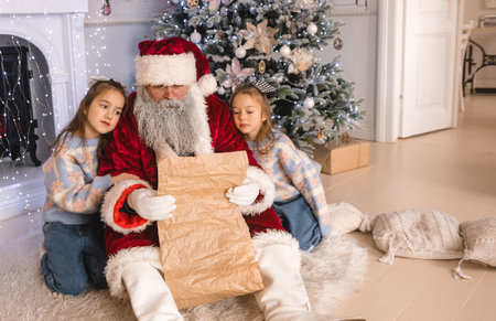 Santa Claus reads a Christmas wish list with two sweet girls nestled beside a decorated Christmas tree. Warm, inviting holiday sceneの写真素材