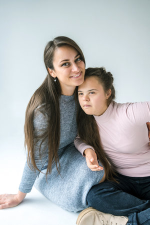 A mother and her daughters with Down syndrome are sitting together and smiling. The mother has her arm around her daughters, and they are both looking at the camera.の写真素材