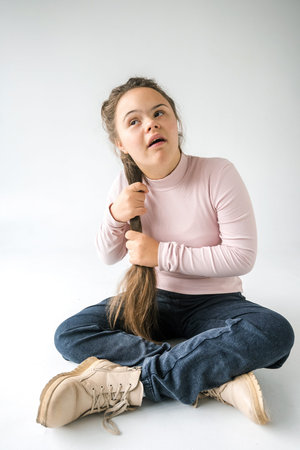 A young woman with Down syndrome sits with her legs crossed, wearing a pink sweater and blue jeans....