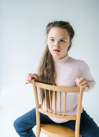 A young woman is sitting on a wooden chair. She is wearing a pink long-sleeved shirt and blue jeans. She is looking up and to the right, with a thoughtful expression on her face. Her long brown hair is falling down her back. She is holding onto the back of the chair with her hands.の写真素材
