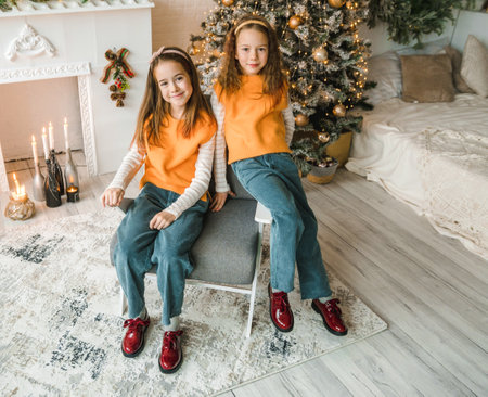 Two adorable twin girls, dressed in matching orange sweaters and denim pants, pose for a Christmas portrait. They are sitting in a chair, wearing bright red shoes, in a room decorated for the holidays.の写真素材