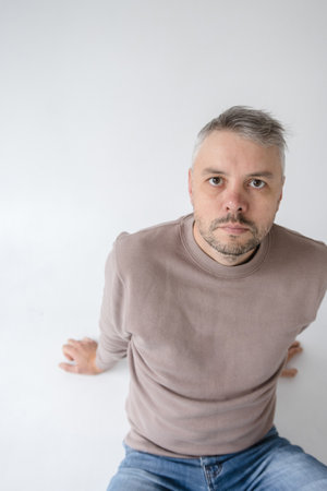 portrait of a contemplative middle-aged man sitting on a white background. He s dressed in a taupe sweatshirt and jeans, his serious expression suggesting thoughtfulness.の写真素材
