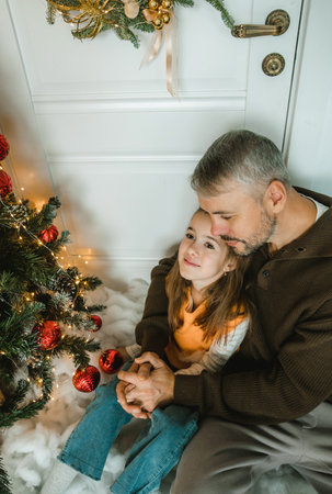 a father and daughter, hands clasped, sharing a tender embrace near a decorated Christmas tree. Warmth and love radiate from the image.の写真素材