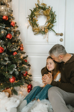 A father and daughter, close and tender, share a heartwarming moment by a Christmas tree. Warmth and love fill the scene.の写真素材