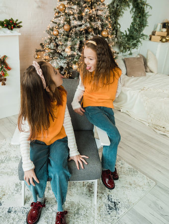 Two playful sisters, dressed in matching orange sweaters and jeans, share a silly moment by the Christmas tree.の写真素材