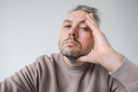 Close-up portrait of a tired or stressed man, hand to his face. His expression conveys fatigue or contemplation. The neutral background emphasizes his emotional state.の写真素材