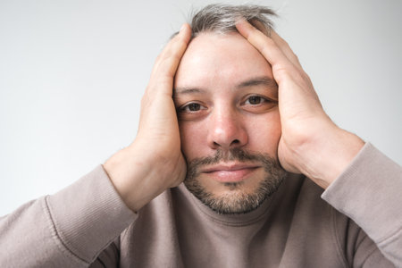 Close-up portrait of a man looking stressed or overwhelmed, his hands covering his face. His expression conveys anxiety, worry, or fatigue.の写真素材