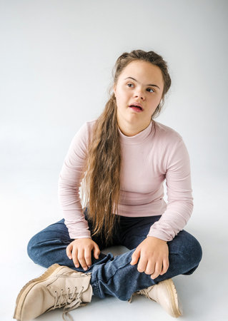 A young woman with Down syndrome sits cross-legged on a white background, looking thoughtfully off to the side. She wears a pale pink long-sleeved top and dark jeans.の写真素材