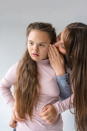 A tender moment between a mother and her teenage daughter with Down syndrome. The mother whispers a secret or a comforting word into her daughter's ear, their closeness conveying a strong bond of love and support.の写真素材