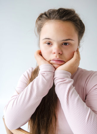 A pensive portrait of a young woman with Down syndrome. She rests her chin in her hands, her...