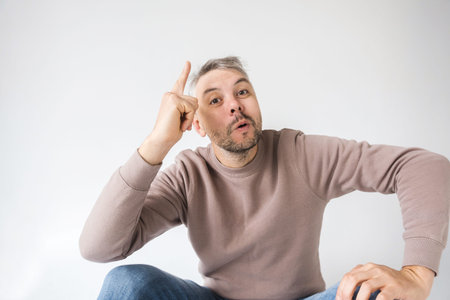 A man sits against a white background, pointing to his head with a playful or insightful expression....