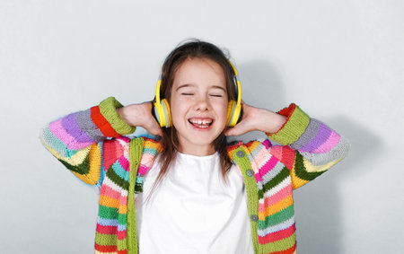 A joyful girl wearing bright yellow headphones and a colorful striped cardigan enjoys listening to music. Her eyes are closed, and she s smiling broadly, expressing pure happiness.の写真素材