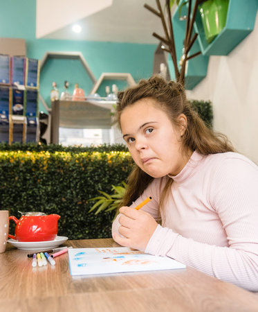 A young woman with Down syndrome sits at a cafe table, appearing thoughtful as she holds a marker...