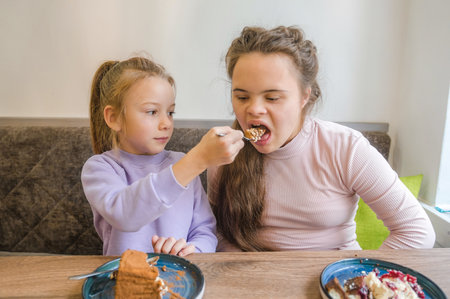 Two young girls are sharing a dessert at a cafe. One girl, who has Down syndrome, is being lovingly fed by her younger sister or friend. The scene is warm, cheerful, and highlights the positive bond between the two girls.の写真素材