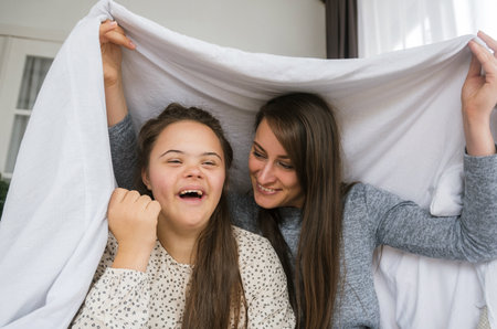 A young woman with Down syndrome and her mother share a joyful, playful moment under a white sheet. They are laughing together, showing a loving and supportive relationship.の写真素材