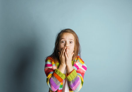A young girl with long brown hair looks shocked or surprised, her hands covering her mouth. She is wearing a brightly colored striped cardigan. The simple light-blue background emphasizes the girl's expression and the vibrant colors of her clothing.の写真素材