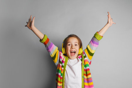 A young girl wearing bright, colorful striped headphones and a matching cardigan joyfully sings or dances with her arms raised. Her open mouth and expressive face show pure happiness and excitement.の写真素材