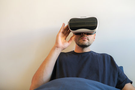 A man uses a VR headset while relaxing in bed. The image focuses on the headset and his relaxed posture, suggesting comfort and immersion in virtual reality.の写真素材