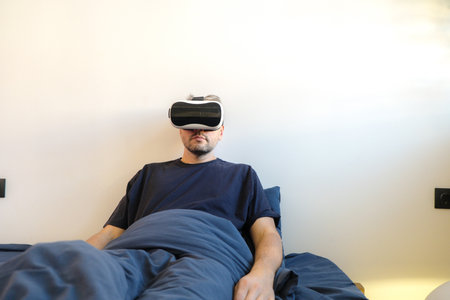 A man relaxes in bed wearing a VR headset. He appears calm and immersed in the virtual world. The scene is peaceful and modern, highlighting the use of VR technology for relaxation and entertainment at home.の写真素材