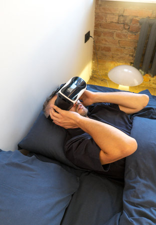 A man lies in bed, comfortably using a VR headset. The setting is a modern bedroom with exposed brick, suggesting a stylish and relaxed atmosphere.の写真素材