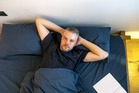 A man lies comfortably in bed, arms behind his head, looking directly at the camera. He s relaxed and at ease under a dark blue duvet. A book rests on the bed beside him.の写真素材