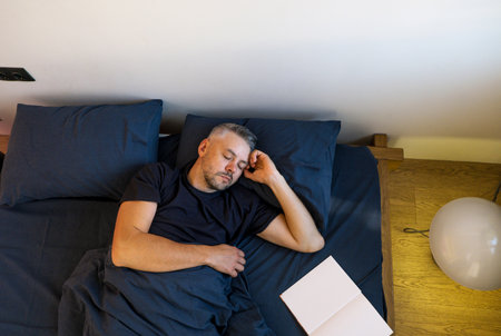 man peacefully sleeping in bed. He s comfortably nestled under a dark blue duvet, with a book resting on the bed beside him. The scene is quiet and serene, perfect for themes of sleep, rest, and relaxation.の写真素材