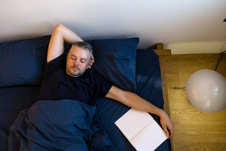 High-angle view of a man sleeping peacefully in bed. He s under a dark blue duvet, one arm resting above his head, the other by his side. A notebook lies open on the bed beside him.の写真素材