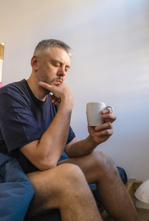 A thoughtful man sits on the edge of his bed, holding a mug and appearing lost in contemplation. The image evokes a sense of quiet reflection and morning introspection.の写真素材