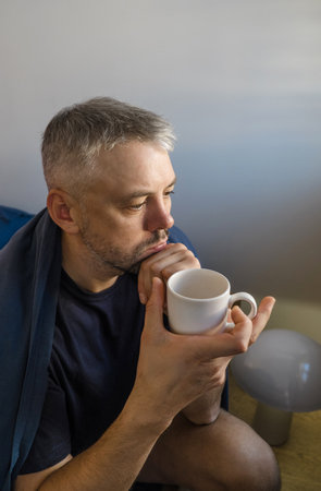 A contemplative man sits on the edge of his bed, holding a warm beverage. He appears lost in thought, creating a mood of quiet reflection and introspection.の写真素材