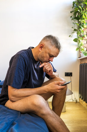 A man sits on his bed, thoughtfully looking at his phone. He appears contemplative, perhaps considering a message or planning his day. The image is casual, suggesting a relaxed morning or downtime at home.の写真素材
