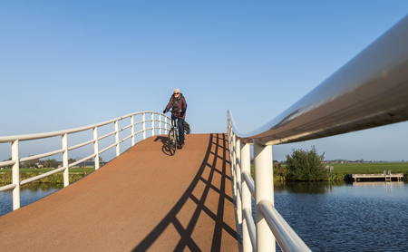 Groot-Ammers, The Netherlands - November 11, 2016: Modern architecture of a cycling bridge over a small canal with three cyclists at Groot-Ammers in the Netherlands.のeditorial素材