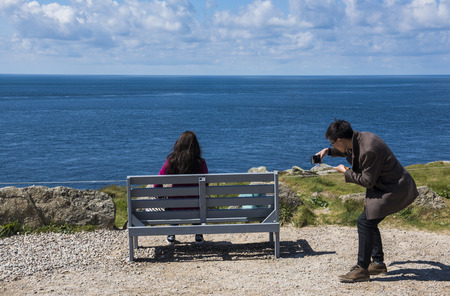 Lands' End, England - April 27, 2017: Tourists sitting on bench at Last/first house at Lands' End in summer taking pictures with phone.のeditorial素材