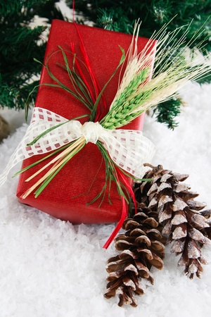 Festive holiday gift wrapped in red recycled paper is decorated with raffia and wheat to create a natural, down-home country feeling, set in a snowy background accented with glittery pinecones and green pine branches.の写真素材