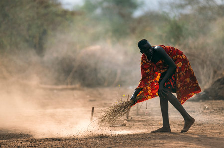 MUNDARI TRIBE, SOUTH SUDAN - MARCH 11, 2020: Man in bright garment sweeping dusty ground with twigs...