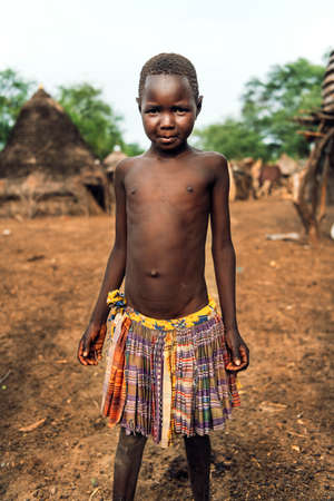 TOPOSA TRIBE, SOUTH SUDAN - MARCH 12, 2020: Boy in colorful skirt looking at camera while living in Toposa Tribe village in South Sudan, Africaのeditorial素材