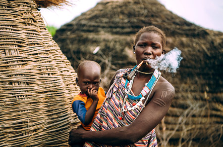 TOPOSA TRIBE, SOUTH SUDAN - MARCH 12, 2020: Woman from Toposa Tribe smoking pipe and carrying baby...