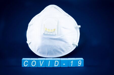 Face mask on dark blue background with covid-19 written on light blue blocks in conceptual image of health threatening virus.の写真素材