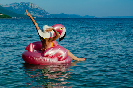 Woman with white straw hat on Flamingo floatie in blue water. Waving a hand, having fun.
