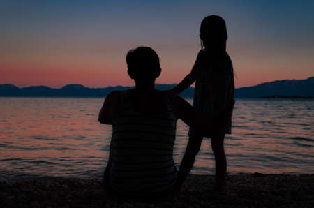 Silhouette of mother sitting on a pebble beach with her daughter standing beside her  watching a beautiful sky after sunset.の写真素材