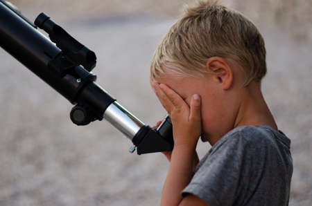 Young boy on a pebble beach looking through a telescope at the evening sky, covering one eye with his hand.の写真素材