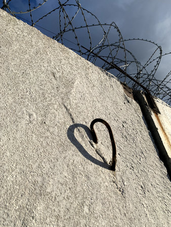 A nail bent into the shape of a heart in a concrete fence topped with barbed wire. Concepts of love in captivity, prison romance, marriage to a prisonerの写真素材