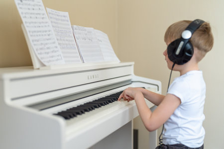 A young boy in a white T-shirt with headphones on his head and learning to play the keys of a white pianoのeditorial素材
