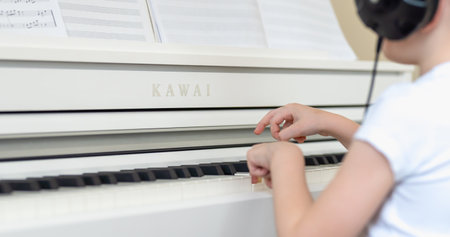 A young boy in a white T-shirt with headphones on his head and learning to play the keys of a white pianoのeditorial素材