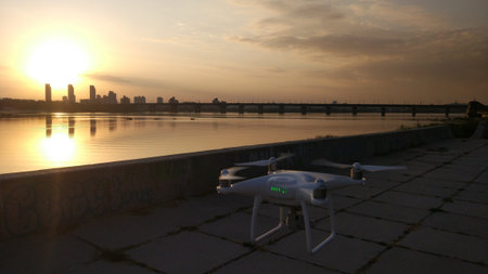 White quadcopter drone fly near the river at the sunset. Cityscape on the background