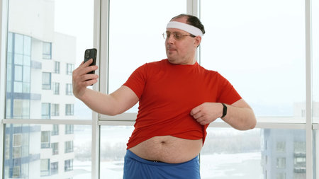 Funny fat man with big belly posing and doing selfie in bright gym or at home. Overweight male...