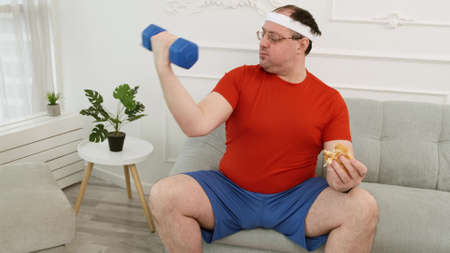 Fat man doing exercise with dumbbells in one hand and eating burger in other hand. Healthy vs unhealthy lifestyle conceptの写真素材