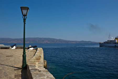 Entry to the harbour on Greek Island of Hydra with Ferry leaving and lamppost in foreground with cannonsの写真素材
