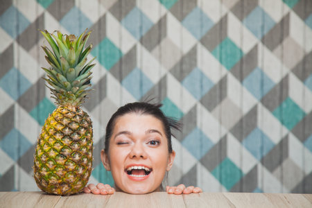 Beautiful girl's head and fresh pineapple on wooden table. Young woman smile and wink. Healthy food...