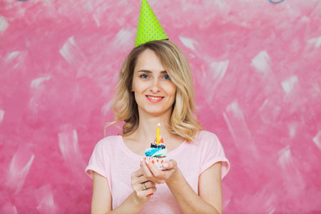 Pretty blonde girl in birthday cap hold cupcake with single candle on a pink background. Colorful holiday cardの写真素材